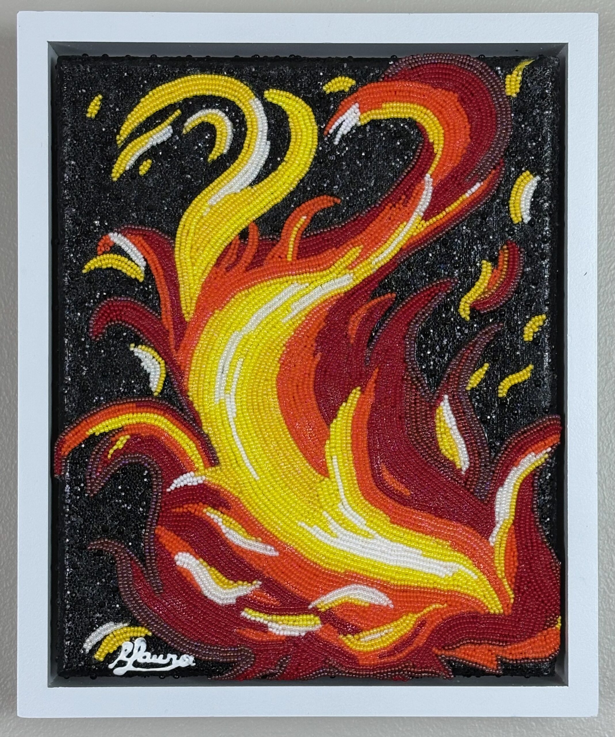 Red, orange, yellow, and white glass beads form expressive flames against a black painted background in Fire, a small bead mosaic in a white frame.